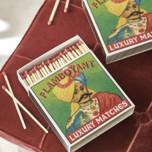May include: Open matchbox with the word "FLAMBOYANT" and "LUXURY MATCHES" printed on the box. The box features an illustration of a man in a turban. The matches are white with red tips. Several loose matches are scattered around the box.