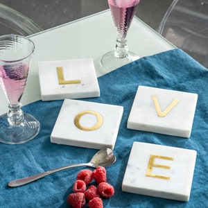 May include: Four white marble coasters with gold letters spelling out the word "LOVE". The coasters are on a blue tablecloth with a spoon and raspberries.