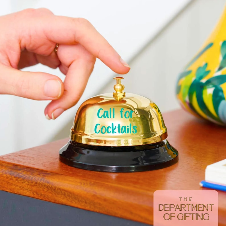 Puede incluir: Una campana dorada con base negra, con el texto "Call for Cocktails" en color turquesa. La campana est&aacute; sobre una superficie de madera, con una mano a punto de tocarla. El logotipo de The Department of Gifting es visible.