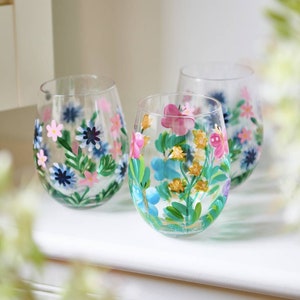 May include: Three clear glass tumblers with hand-painted floral designs. The designs feature pink, blue, yellow, and green flowers.