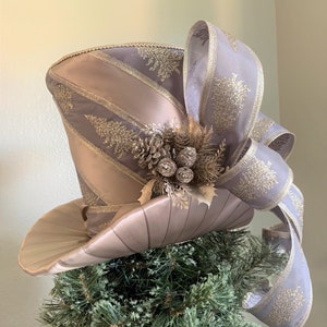 May include: A decorative top hat tree topper in shades of champagne and gray, adorned with gold accents. The hat features a wide brim and is embellished with a large bow and decorative pinecones. The topper is designed to sit atop a Christmas tree.