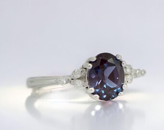 May include: A silver ring featuring an oval, dark blue gemstone as the centerpiece. The ring has a delicate design with smaller, clear stones on either side of the main stone, adding sparkle. The band is thin and polished, reflecting light.