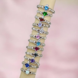 May include: A collection of silver-toned Claddagh rings, each featuring a heart-shaped gemstone in various colors, including blue, green, red, and purple. The rings are displayed on a light-colored cylindrical stand against a soft pink background.