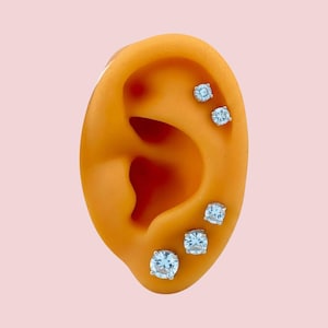 May include: Five silver stud earrings with square, light blue gemstones are displayed on a prosthetic ear. The ear is a warm orange color, set against a soft pink background. The earrings are evenly spaced on the ear.