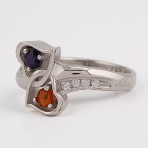 May include: A silver ring with a unique design. The ring features two heart-shaped settings, one holding a dark purple gemstone and the other an orange gemstone. A row of small, clear stones is set into the band. The name "Alex" is engraved on the side.