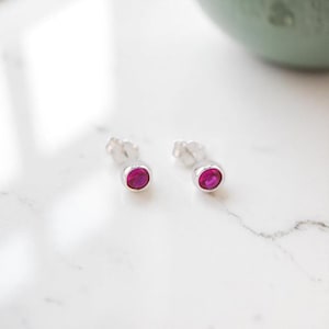 May include: A pair of silver stud earrings with round, bright pink gemstones. The earrings are set in a simple bezel setting, and the posts are visible. The earrings are displayed on a white marble surface.