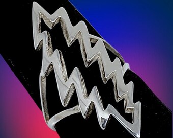 May include: A silver ring featuring a zig-zag pattern. The ring's design is modern and geometric, with multiple layers of the zig-zag motif. It's presented on a black surface, set against a gradient background of blue, purple, and red.