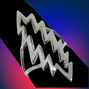 May include: A silver-colored ring with a zig-zag design. The ring has a modern, geometric shape, with multiple layers of the zig-zag pattern. The ring is displayed on a black surface against a gradient background of blue, purple, and red.