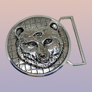 May include: A silver-colored belt buckle featuring a detailed bear head with three eyes. The bear is centered on a circular plate with a dotted grid pattern. The buckle has a rectangular bar for the belt.