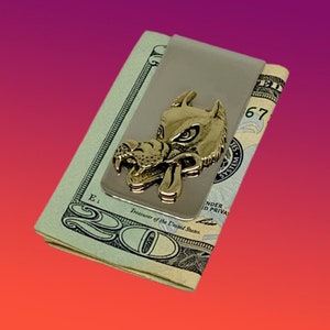 May include: A silver money clip with a gold-toned wolf head design. The wolf's mouth is open and its eyes are fierce. The money clip is holding a twenty dollar bill.