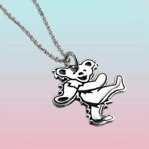 May include: A silver-colored pendant necklace featuring a dancing bear design. The bear is in a stylized, cartoonish form, with a smiling face and outstretched arms and legs. The necklace has a delicate chain.