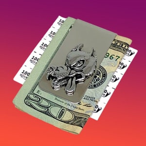 May include: Silver money clip with a wolf head design. The wolf has a snarling expression and a long tongue. The money clip is holding a stack of US dollar bills.