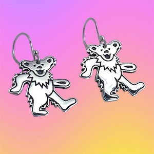 May include: A pair of silver-colored bear-shaped earrings. Each earring features a stylized bear with outstretched arms and legs, with a jagged collar. The earrings hang from a simple silver hook. The background is a gradient of pink, purple, and yellow.