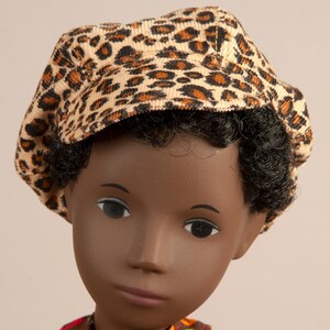May include: A doll wearing a tan corduroy cap with a leopard print pattern. The cap is brown and black. The doll has dark skin, black curly hair, and is wearing a patterned shirt.