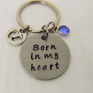 May include: Silver keychain with a blue rhinestone charm and a charm with a footprint design. The main charm is a silver circle with the text "Born in my heart".