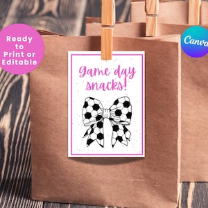 May include: A printable gift tag with a pink border and the text "game day snacks!" in pink lettering. A black and white soccer ball bow is in the center of the tag.