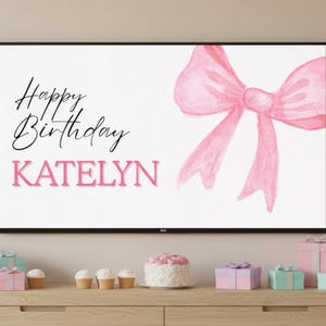 May include: A white background with a pink watercolor bow and the words "Happy Birthday KATELYN" in black and pink. The image is displayed on a black framed screen. Below the screen are wrapped gifts, cupcakes, and a cake.