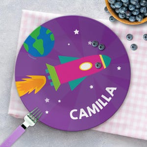 May include: A purple plate with a rocket ship design, the word "CAMILLA" in white, and a cartoon Earth. The rocket is pink, green, and blue with an orange flame. White stars are scattered around the design.