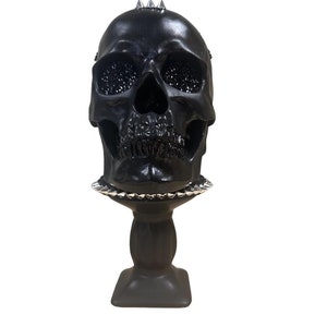May include: A black skull sculpture with silver accents, including a crown of spikes and a decorative base. The skull's eye sockets are filled with a textured material. The sculpture is a gothic or Halloween-themed decorative item.