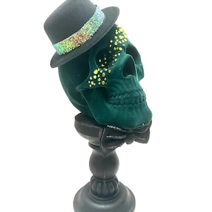 May include: A decorative green skull with a black top hat featuring a shimmering iridescent band. The skull has gold-colored embellishments and a black bow tie. It is mounted on a black pedestal.