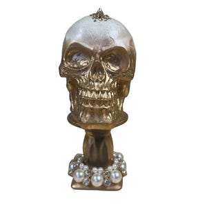 May include: A decorative skull figurine with a gold finish. The skull has a white and gold top, with a gold base and a pearl and rhinestone necklace. The figurine is a unique home decor item.