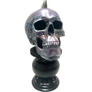 May include: A decorative skull sculpture with a metallic silver finish and iridescent accents. The skull features a row of metal spikes on top and is adorned with colorful gems. It sits atop a black pedestal with additional spikes.