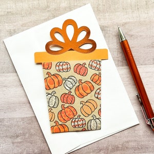May include: A white card with a brown and orange gift box design. The gift box is decorated with a pattern of orange and brown pumpkins. The card is blank and ready for a message.