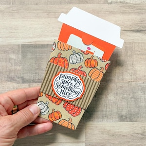 May include: A coffee cup-shaped gift card holder featuring a tan background with a repeating orange and black pumpkin pattern. A brown corrugated paper sleeve wraps around the center with a white label that reads pumpkin spice & something nice.