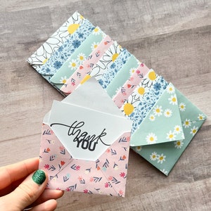 May include: A set of small, colorful envelopes with floral patterns. The envelopes are folded and stacked on top of each other. The top envelope is open and has a white card inside with the words "thank you" written in black ink.