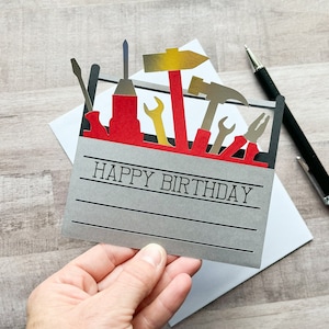 May include: A gray card with the words "Happy Birthday" printed on it. The card features a red toolbox with various tools, including a hammer, screwdriver, and wrenches.