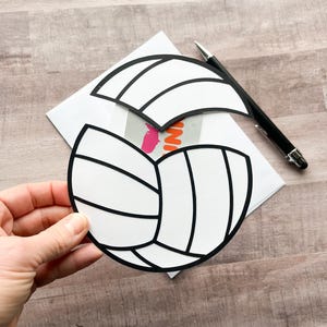 May include: A white and black volleyball card with a cut-out shape for a gift card or message. The card is on a wooden surface.