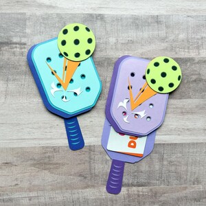 May include: Two papercraft paddleball rackets with a green ball on top. The rackets are purple and blue with orange accents. The rackets are folded to reveal a card inside.