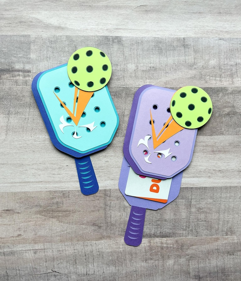 May include: Two papercraft paddleball rackets with a green ball on top. The rackets are purple and blue with orange accents. The rackets are folded to reveal a card inside.