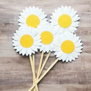May include: Five white paper daisy flowers with yellow centers on wooden sticks. The flowers are arranged in a bouquet.