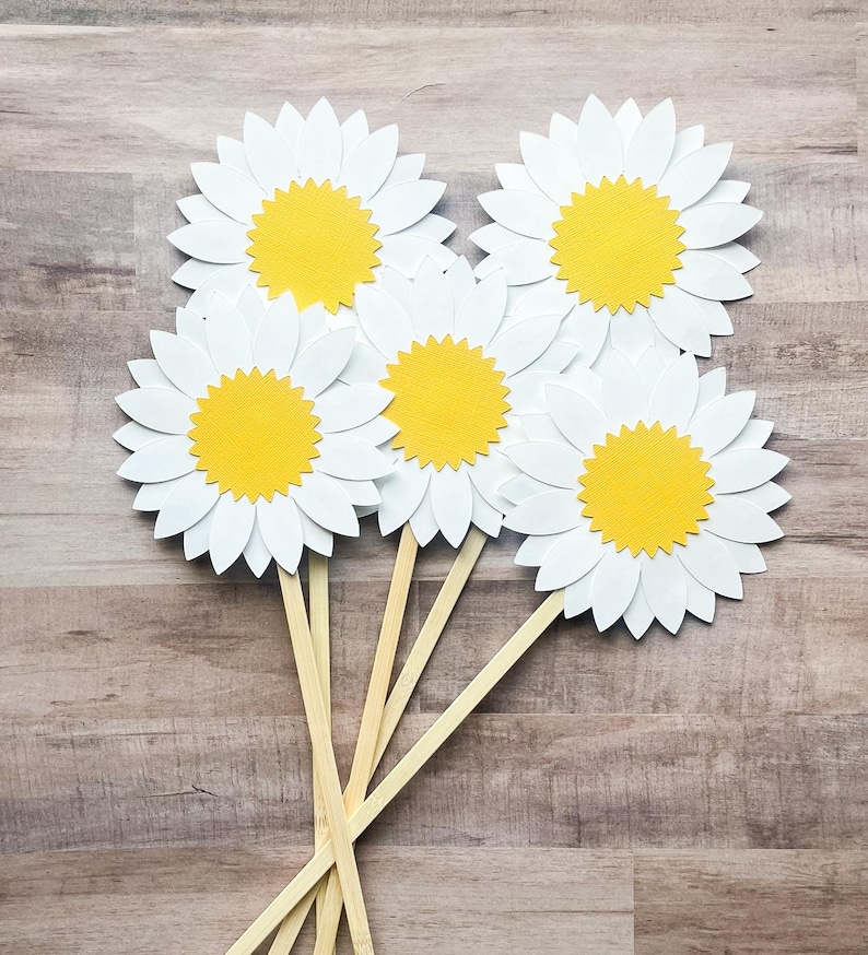 May include: Five white paper daisy flowers with yellow centers on wooden sticks. The flowers are arranged in a bouquet.
