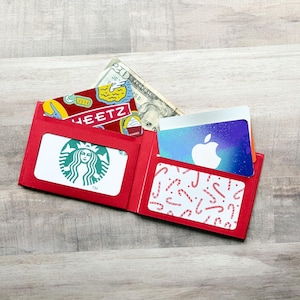 May include: A bright red bi-fold wallet featuring two clear plastic windows and an interior pocket. One window displays a green and white Starbucks logo card, while the other shows a candy cane pattern. A twenty-dollar bill and an Apple logo card are visible.