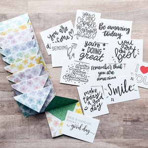 May include: A collection of white cards with black handwritten motivational phrases, such as "Be amazing today" and "Smile." The cards are accompanied by small, floral-patterned envelopes in pastel colors. The envelopes are arranged in a row.