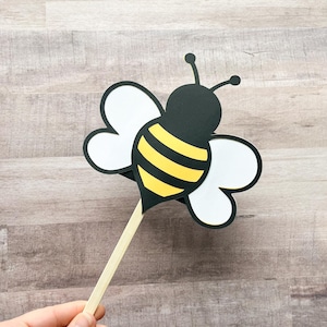 May include: A black and yellow bee with white wings on a wooden stick. The bee is a cutout and is used as a prop for photos.