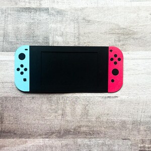 Peut inclure: Une console de jeu noire avec un &eacute;cran et des manettes bleues et rouges. Les manettes ont des boutons noirs. La console est pos&eacute;e sur une surface en bois.