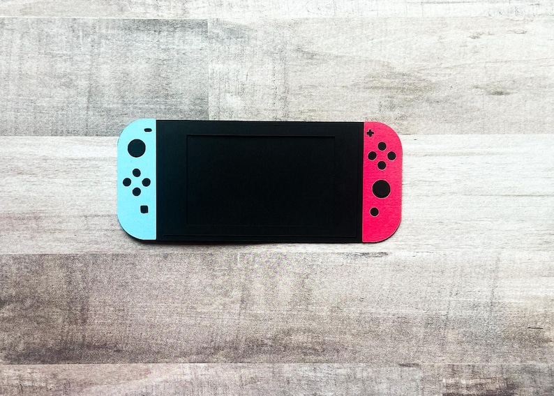 Peut inclure: Une console de jeu noire avec un &eacute;cran et des manettes bleues et rouges. Les manettes ont des boutons noirs. La console est pos&eacute;e sur une surface en bois.