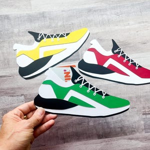 May include: Three colorful paper cut-out sneakers in yellow, red, and green. Each shoe has a white sole, black accents, and white laces. The green shoe is being held by a hand.