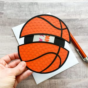 May include: A card with a pop-up basketball design. The basketball is orange with black lines and a textured surface. The card is white.
