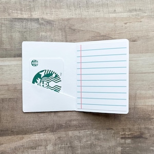 May include: A white card holder with a Starbucks gift card in the left pocket. The right side has lined paper with a pink vertical line. The Starbucks card has a green logo.