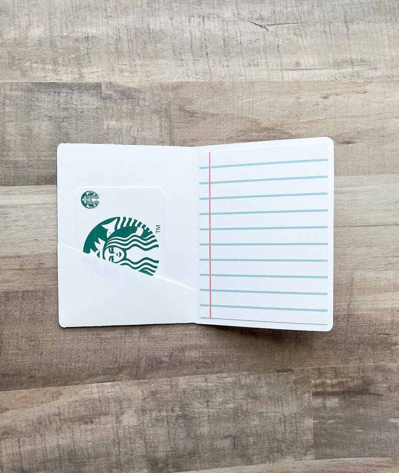 May include: A white card holder with a Starbucks gift card in the left pocket. The right side has lined paper with a pink vertical line. The Starbucks card has a green logo.