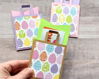 May include: Three Easter-themed gift bags made of paper with a handle. The bags are decorated with a pattern of colorful Easter eggs. The bags are folded flat and ready to be assembled.