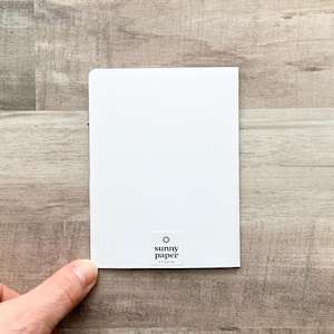 May include: A white card with rounded corners, featuring the "sunny paper studio" logo in the bottom right corner. The card is held by a hand against a light brown wooden surface.