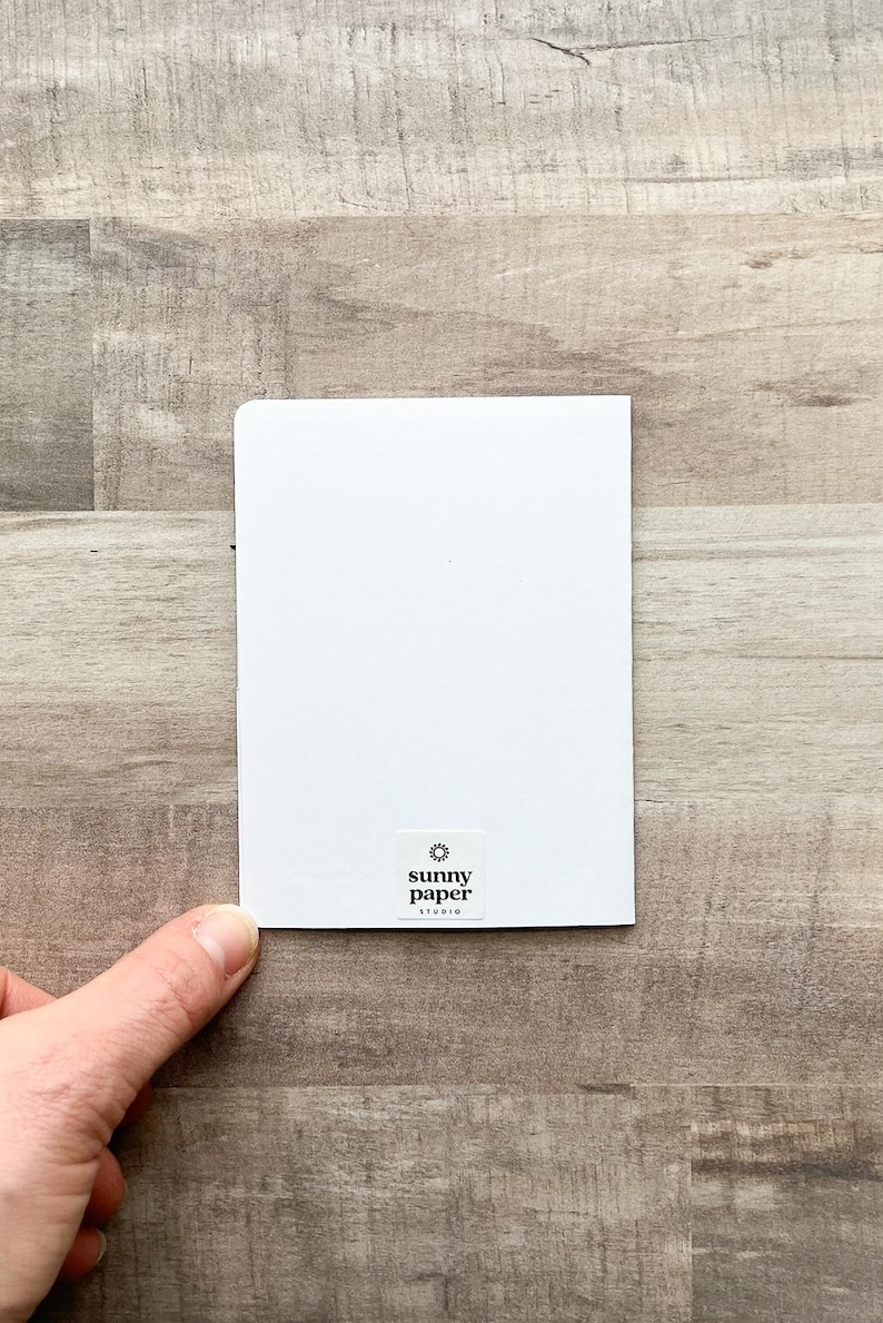 May include: A white card with rounded corners, featuring the "sunny paper studio" logo in the bottom right corner. The card is held by a hand against a light brown wooden surface.
