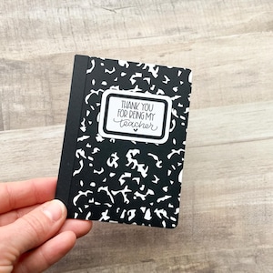 May include: A small black and white composition notebook with the text "THANK YOU FOR BEING MY TEACHER" in a white rectangular frame. The notebook is held in a hand and has a black spine.
