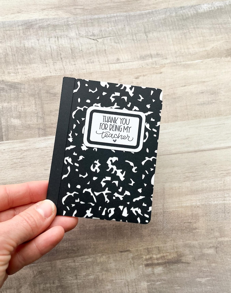 May include: A small black and white composition notebook with the text "THANK YOU FOR BEING MY TEACHER" in a white rectangular frame. The notebook is held in a hand and has a black spine.