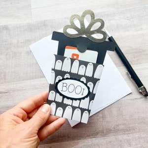 May include: A Halloween-themed gift card holder with a black and white ghost pattern. The holder has a black top with a silver decorative element and the word "BOO!" on the front. A pen and envelope are in the background.